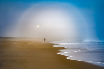 Person walking on sandy beach with sun through hazy mist and  clouds with ice rainbow and waves