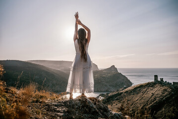 A woman stands on a hill overlooking the ocean. She is wearing a white dress and has her hands on...