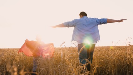 Father and son superhero in red cloak flying running at sunny dry wheat field back view. Happy...