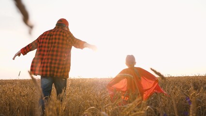 Father farmer and little daughter superhero in red cloak running at sunset wheat field. Happy...