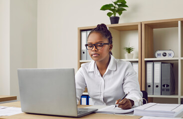 Serious, focused young, beautiful africanamerican girl with glasses, dressed in a white shirt, is...