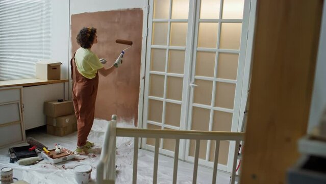 Back full footage of single mother in late pregnancy stage painting walls with roller in brown while remodeling room into nursery for future child