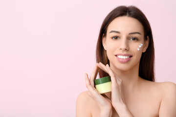 Beautiful young woman with cream on cheek holding jar of sunscreen against pink background