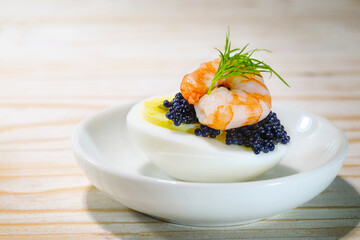 Halved boiled eggs with black caviar, prawn and dill garnish on a small white plate and a wooden table, finger food snack for holidays like Easter, Christmas or New Year, copy space