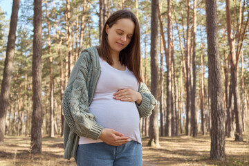 Beautiful pregnant woman wearing green jumper stroking her belly with hands smiling with love...