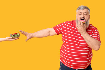 Overweight mature man unhealthy donut refusing healthy vegetable salad on yellow background. Weight...