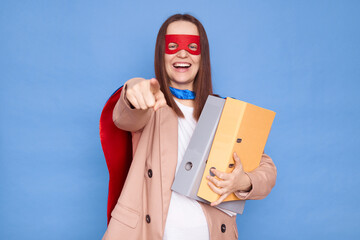 Excited woman wearing superhero costume and mask holding folders isolated over blue background...