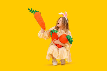 Beautiful young shocked woman in bunny ears with carrot-shaped toys on yellow background. Easter celebration
