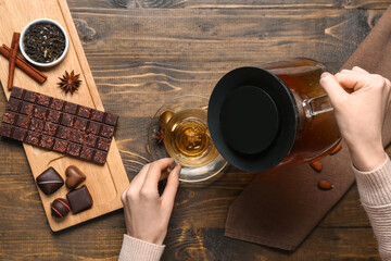 Woman pouring tasty hot tea from teapot into cup and tray with chocolate on wooden background - Powered by Adobe