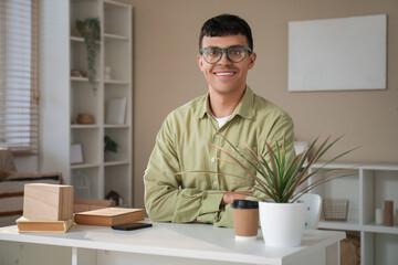 Handsome happy young man with new stylish eyeglasses sitting at table in room