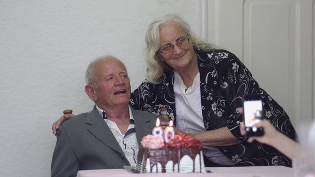 A man with his wife sits in front of a cake with candles on it celebrating 80th birthday - Powered by Adobe