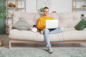 Male student with piggy bank and laptop on sofa in room. Concept of savings for education