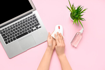 Woman with laptop and bottle of sanitizer wiping mouse on pink background