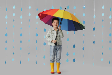 Cute little Asian boy in gumboots with umbrella and paper raindrops on white background