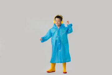 Cute little Asian boy in raincoat with gumboots and headphones on white background