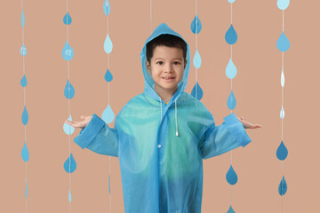 Cute little Asian boy in raincoat with paper raindrops on beige background