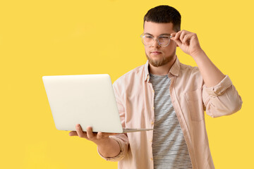 Young man in eyeglasses using laptop on yellow background