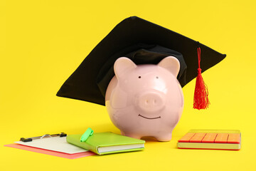 Piggy bank in graduation hat with stationery on yellow background