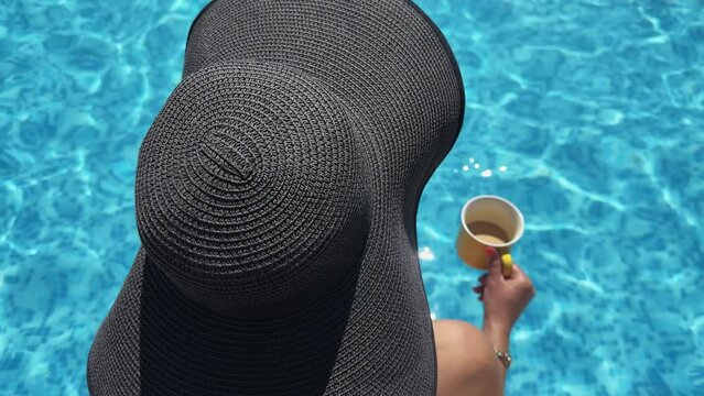 A woman wearing a black hat is sitting by a pool with a cup of coffee in her hand