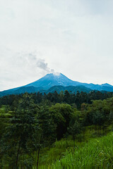 Mount Merapi in Indonesia in the afternoon is very clear and majestic against a background of blue sky and green forest with a ravine flowing cold lava in the foreground.