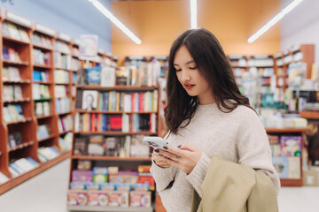 Serious teen gen Z Korean student girl holding mobile phone, using app on smartphone in college library, browsing, chatting, consulting internet, getting cheerful, happy, smiling. Part of a series