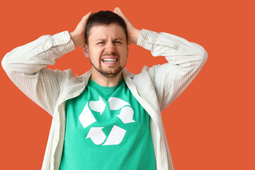 Angry young man in t-shirt with recycling logo holding head on orange background. Ecology concept