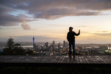 Tourist taking selfies using smartphone. Sky Tower and cityscape in the background. Mt Eden summit at dawn. Auckland.
