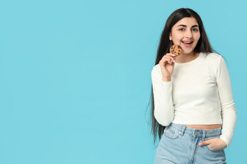 Beautiful young happy woman holding tasty cookie with chocolate chips on blue background