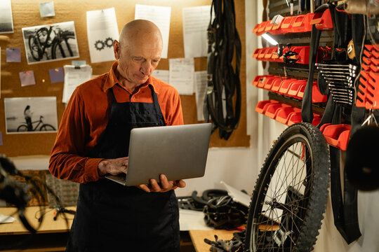 Mature Repairman using laptop in his garage. Male mechanic Standing In His Workshop. Elderly Bike shop owner with laptop computer
