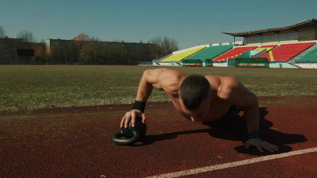 Young man training at city stadium and performs push-ups from kettlebell rearranging hands. Slow motion. Challenging exercise that works chest, triceps and core muscles