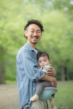 新緑の屋外で赤ちゃんを抱くパパ　イクメンや男性の育児や子育てのイメージ　カメラ目線の縦