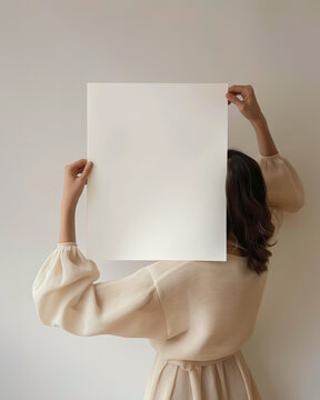 A woman female artist with dark hair with hands holding a blank 16 x 20, 40 x 50 art print mockup, in a minimal neutral interior, the art print covers her face
