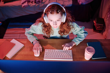 From above view shot of red-haired girl wearing headphones playing online video game on desktop...