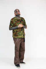 Full body shot of an asian man with full face tattoos, eye tattoo, flesh tunnels, and septum, nose bridge and upper lip piercings. Wearing a camouflage shirt and brown pants