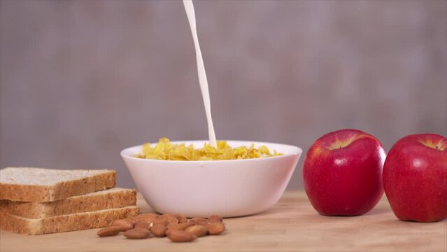 Pouring milk in a bowl containing cornflakes. Red apple and whole wheat breads on the table. Slow motion 4K.