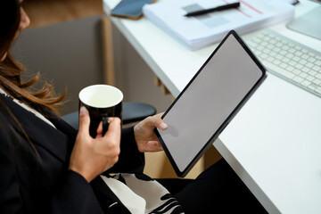 Close up shot of young businesswoman holding cup of coffee using digital tablet at her office desk