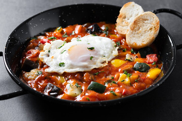 Vegetable pisto manchego with tomatoes, zucchini, peppers, onions,eggplant and egg, served in frying pan on black slate background