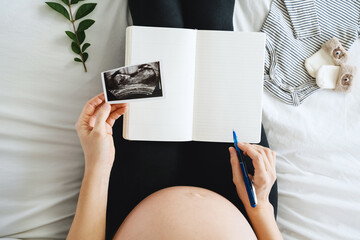 Pregnant woman with blank planner notebook, ultrasound images and clothes for baby.