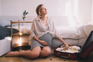 Pregnant woman is packing suitcase for maternity hospital getting ready for childbirth. Happy young mother with travel luggage of baby clothes at home.