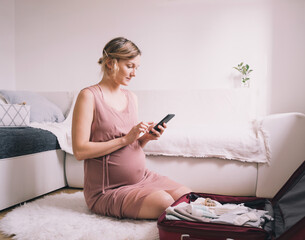Pregnant woman packing suitcase for hospital or journey, getting ready for newborn baby, labor, making list or taking photo with smartphone. Clean clothes and things in bag.