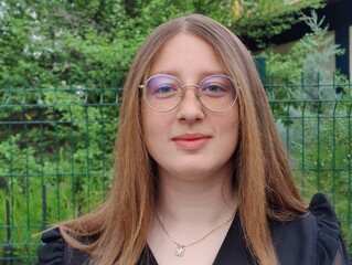 The image shows a person standing in front of a green fence with foliage behind it. The person has long, straight hair and is wearing glasses with a round frame. Portrait with a focus on the person.