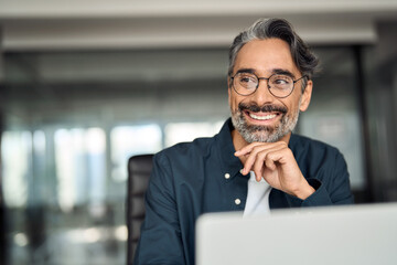 Smiling mature business man executive wearing shirt sitting at desk using laptop. Happy busy professional middle aged Indian businessman investor working on computer looking away in office. Copy space