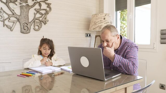 Girl coloring with her grandpa while working on a laptop at home
