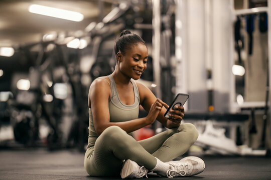 A fit african american sportswoman is typing messages on her phone while sitting on a gym floor.