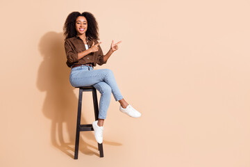 Full length photo of nice girl wear brown blouse jeans sit on stool indicating at logo empty space...
