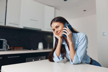 Woman sitting on kitchen counter talking on cell phone while looking at camera in casual home setting