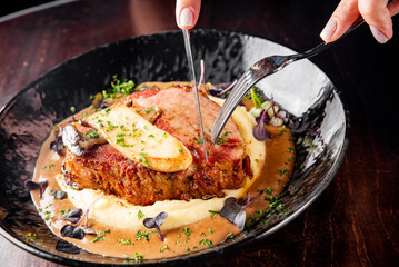 Person cutting a succulent steak topped with butter, served on mashed potatoes, garnished with herbs in a black bowl