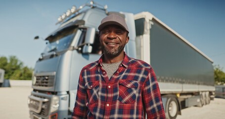 close up of young smiling truck driver standing outside on background of truck cars. Happy african...
