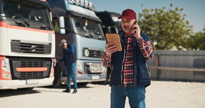 Adult Caucasian trucker speaking on his phone with his coworkers or customers about delivery planning. Busy man actively communicating in front of lorries. Shipping process. Transportation industry.