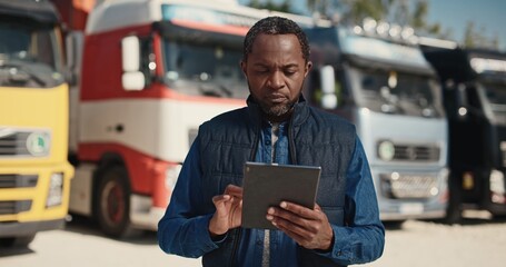 Attractive African American male driver using his work tablet device while standing before trucks....